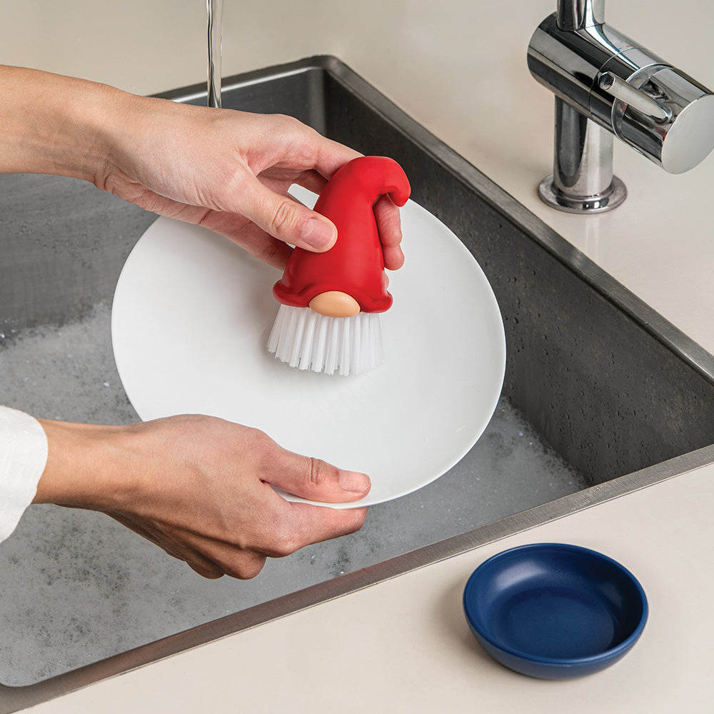 Person using a red gnome-shaped scrubber to clean a white plate in a kitchen sink